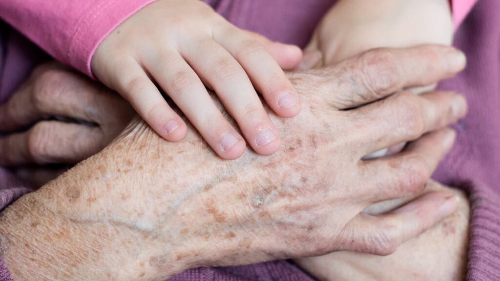 Gros plan sur les mains d'un enfant dans celles d'une personne âgée illustrant l'apparition de taches de vieillesse sur les mains